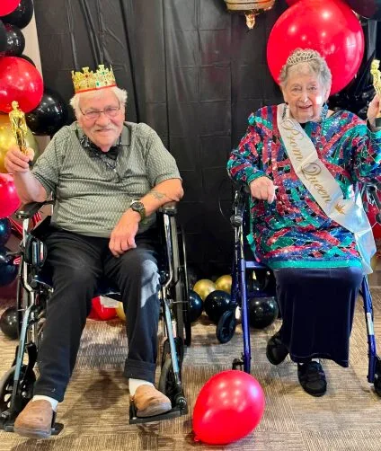 Senior prom king and queen in their sash and crown surrounded by balloons | Aspen Creek Senior Living